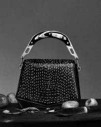 Black handbag with white kantha stitching and a curved handle resting on a dark surface alongside potatoes and a knife.