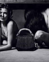 Black and white fashion campaign image featuring a structured handbag with a curved handle and light stitching, positioned before two seated people.