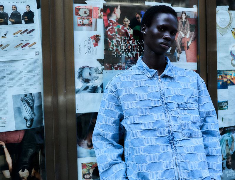 Portrait of a Black adult male wearing a patterned denim jacket, standing against a wall covered with magazine clippings.