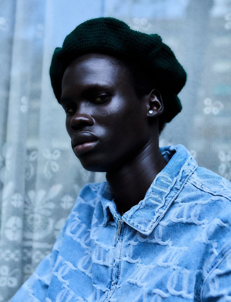 Portrait of adult male with dark skin wearing a knit beret and textured denim jacket in front of a blurred window with sheer curtains.
