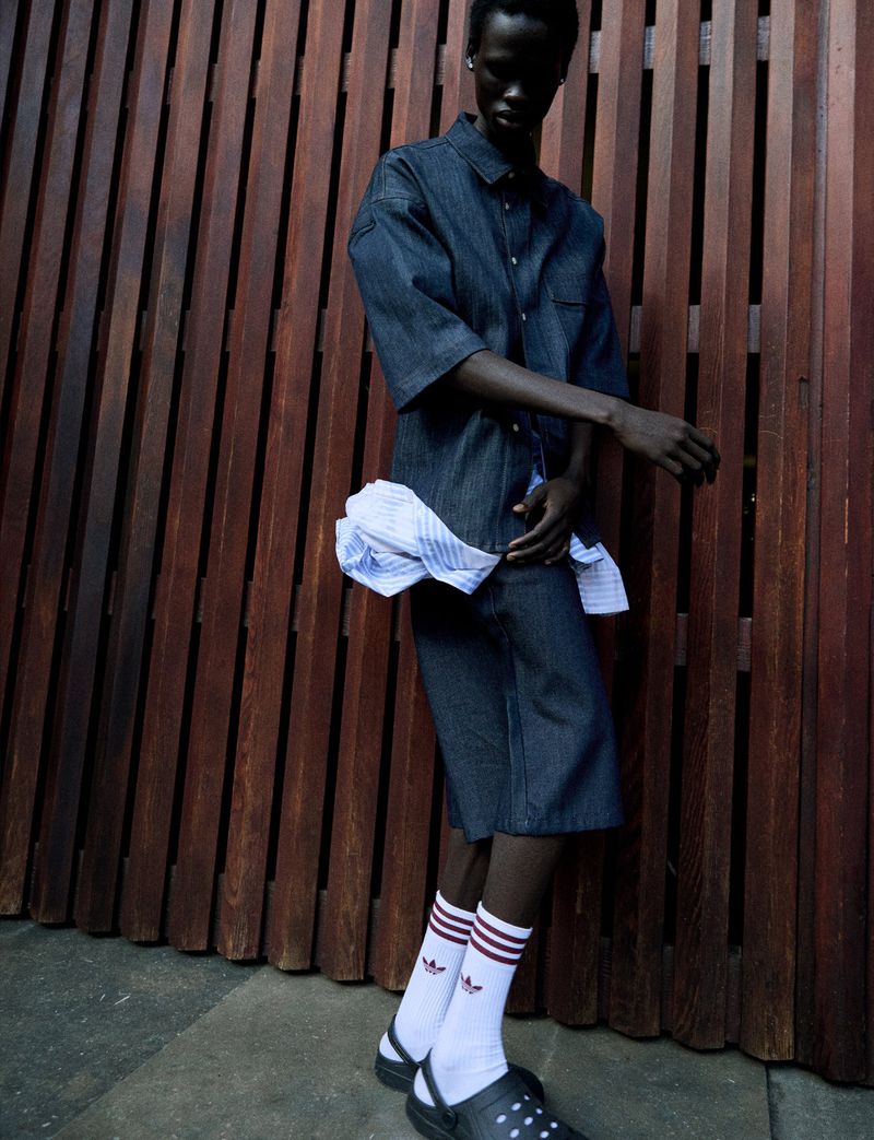 A person in a blue denim outfit with white socks and black clogs, posing against a brown wooden wall, fashion editorial style.