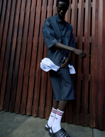 A person in a blue denim outfit with white socks and black clogs, posing against a brown wooden wall, fashion editorial style.