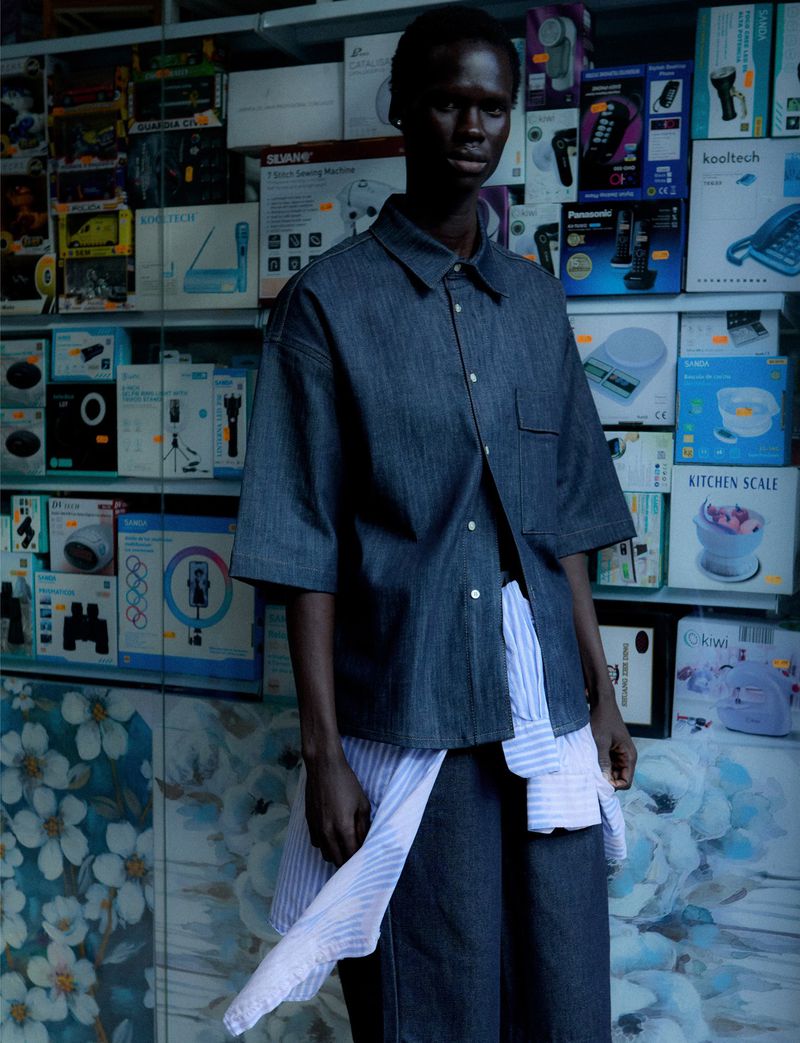A young woman poses in a casual denim outfit with a blue and white striped shirt, standing in front of stacked electronic product boxes.