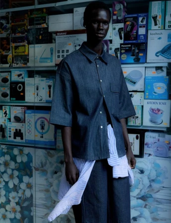 A young woman poses in a casual denim outfit with a blue and white striped shirt, standing in front of stacked electronic product boxes.
