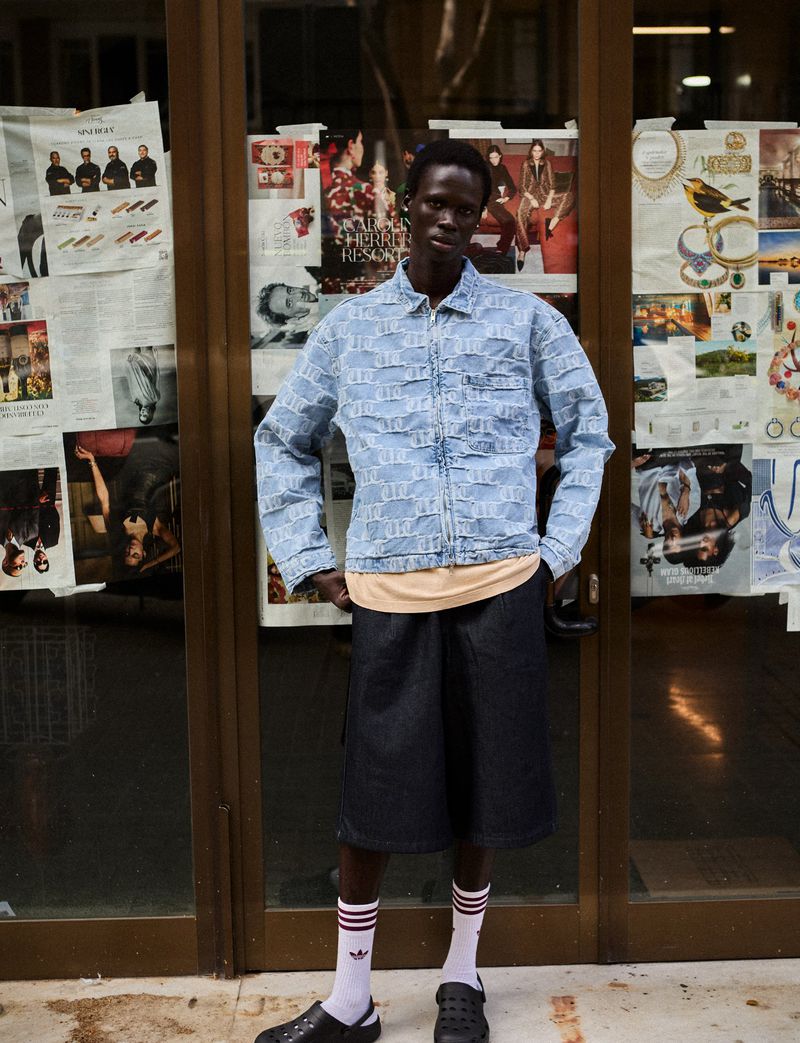 An adult male stands in front of a glass door plastered with posters, wearing a light blue jacket, dark shorts, white socks, and black clogs.