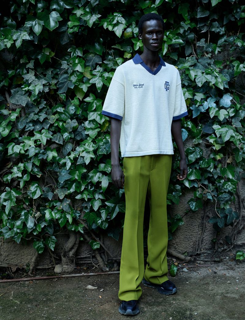 Fashionable adult male model in an ivory polo shirt and olive-green pants stands against a lush ivy wall outdoors.
