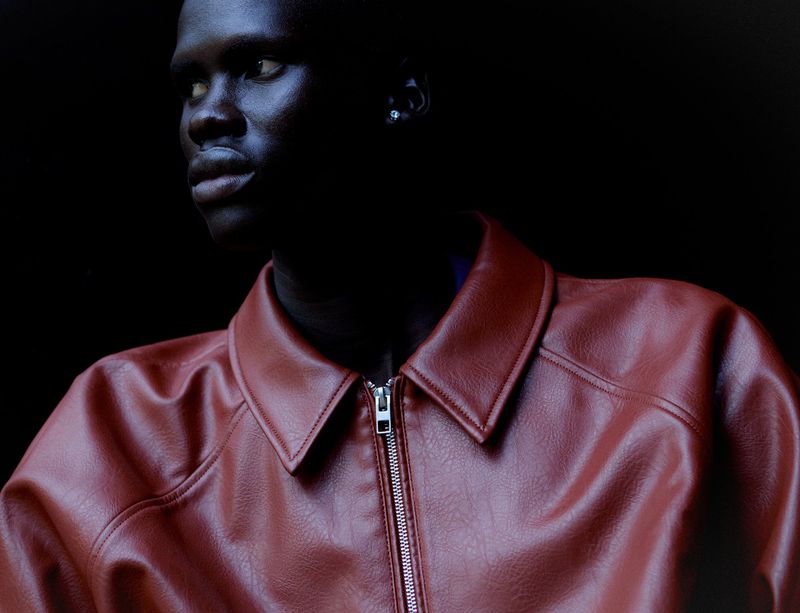 Head and shoulders portrait of a dark-skinned adult male wearing a reddish-brown leather jacket against a dark background, looking left.