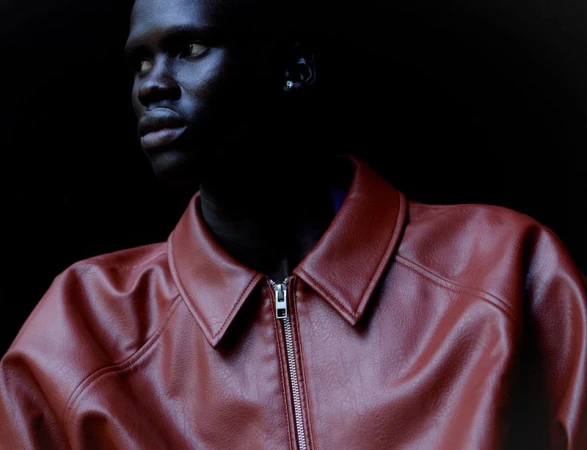 Head and shoulders portrait of a dark-skinned adult male wearing a reddish-brown leather jacket against a dark background, looking left.
