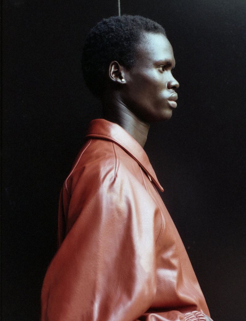 Profile portrait of an adult male with dark skin wearing a brown leather jacket against a dark neutral backdrop in a studio setting.