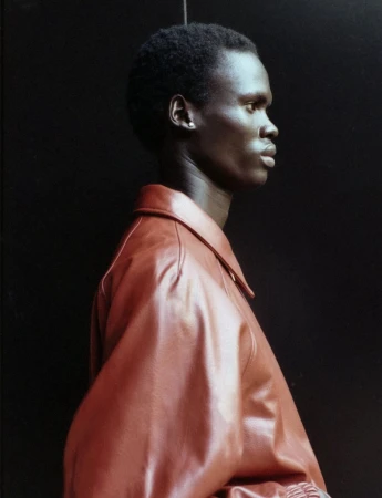 Profile portrait of an adult male with dark skin wearing a brown leather jacket against a dark neutral backdrop in a studio setting.