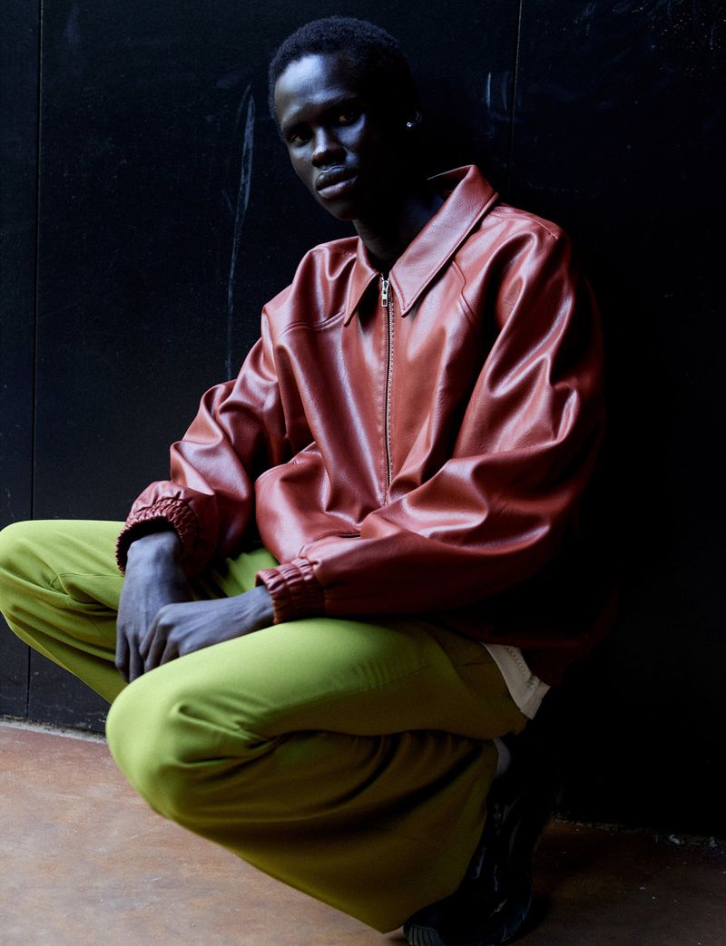 A person in a brown leather jacket and green pants crouches against a dark wall, looking at the camera in a fashion style portrait.