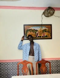 A shirtless black man wearing a blue patterned jacket and beige trousers standing indoors against an off-white wall with a framed painting.