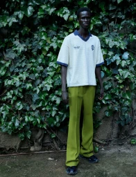 Model standing outdoors wearing a white knit polo shirt and green trousers against a wall of lush ivy leaves