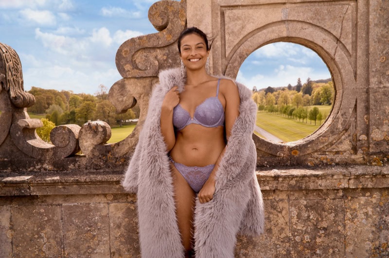 Model standing outdoors wearing purple lace lingerie and a fluffy grey coat against stone architecture