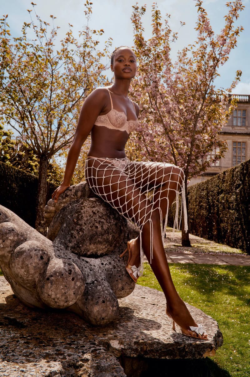 Model seated outdoors on stone sculpture wearing pink lingerie set and white net skirt beneath blossoming trees.