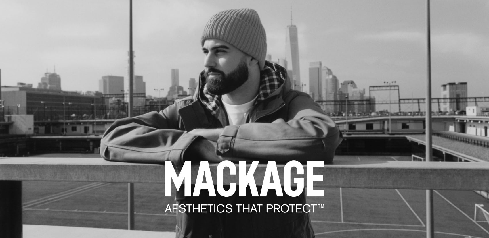 Man wearing a beanie and winter jacket leaning against a railing with city skyline in the background for Mackage Fall 2024 ad campaign