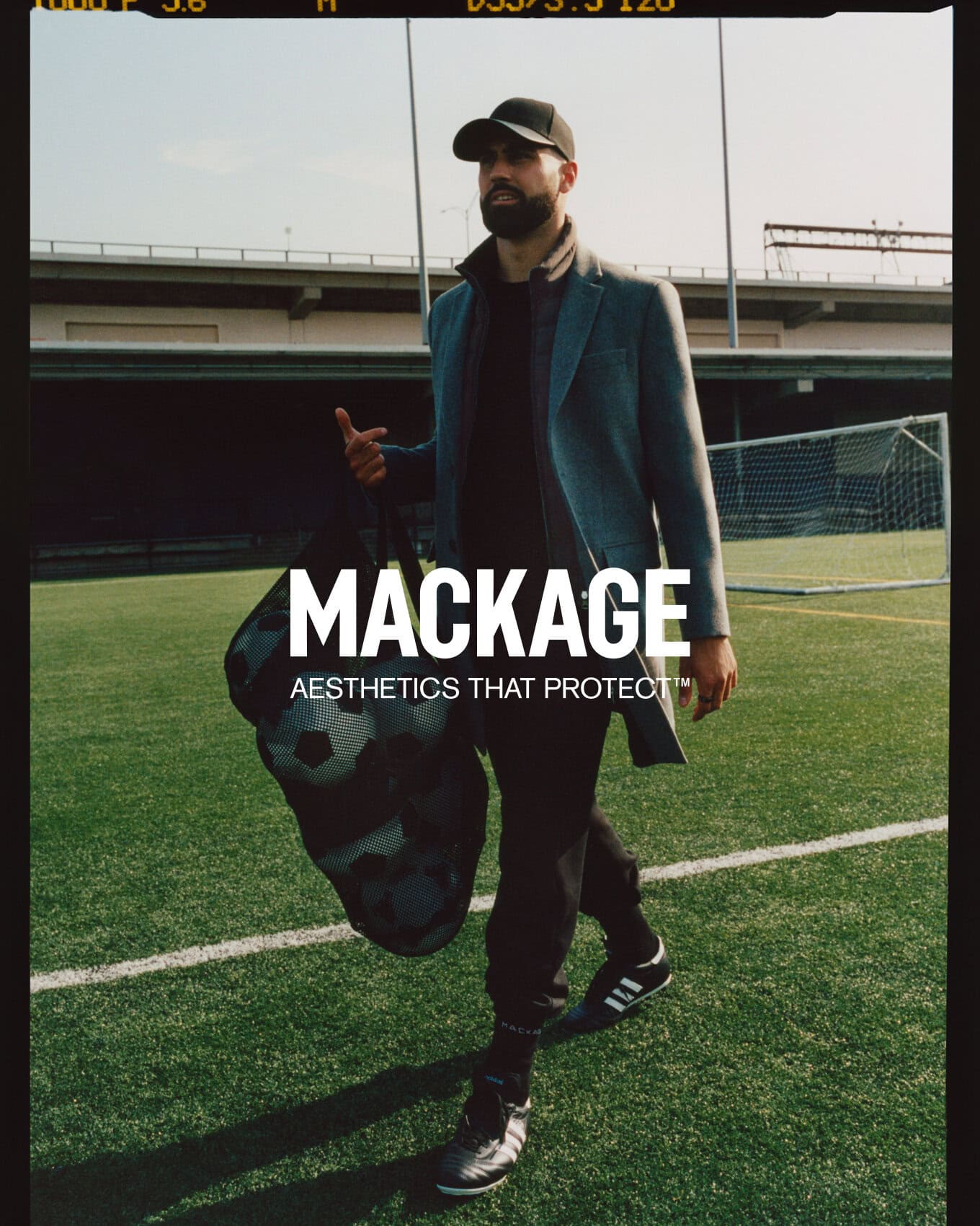 Man wearing a grey coat and baseball cap walking across a green sports pitch carrying a mesh bag filled with soccer balls