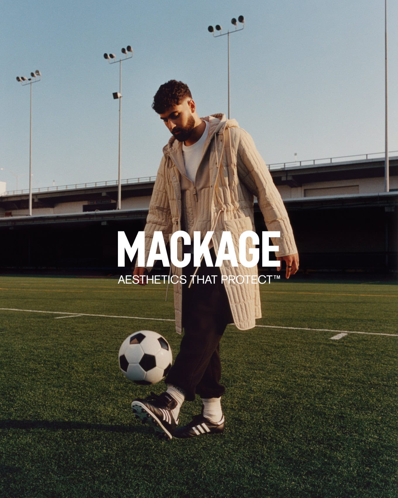 Man wearing a quilted hooded coat juggling a soccer ball on a turf field with stadium lights and a highway overpass behind him.