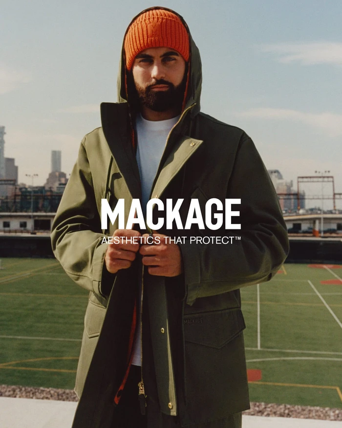 Man wearing a hooded olive green jacket and bright orange beanie standing on a rooftop sports court