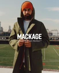 Man wearing a hooded olive green jacket and bright orange beanie standing on a rooftop sports court