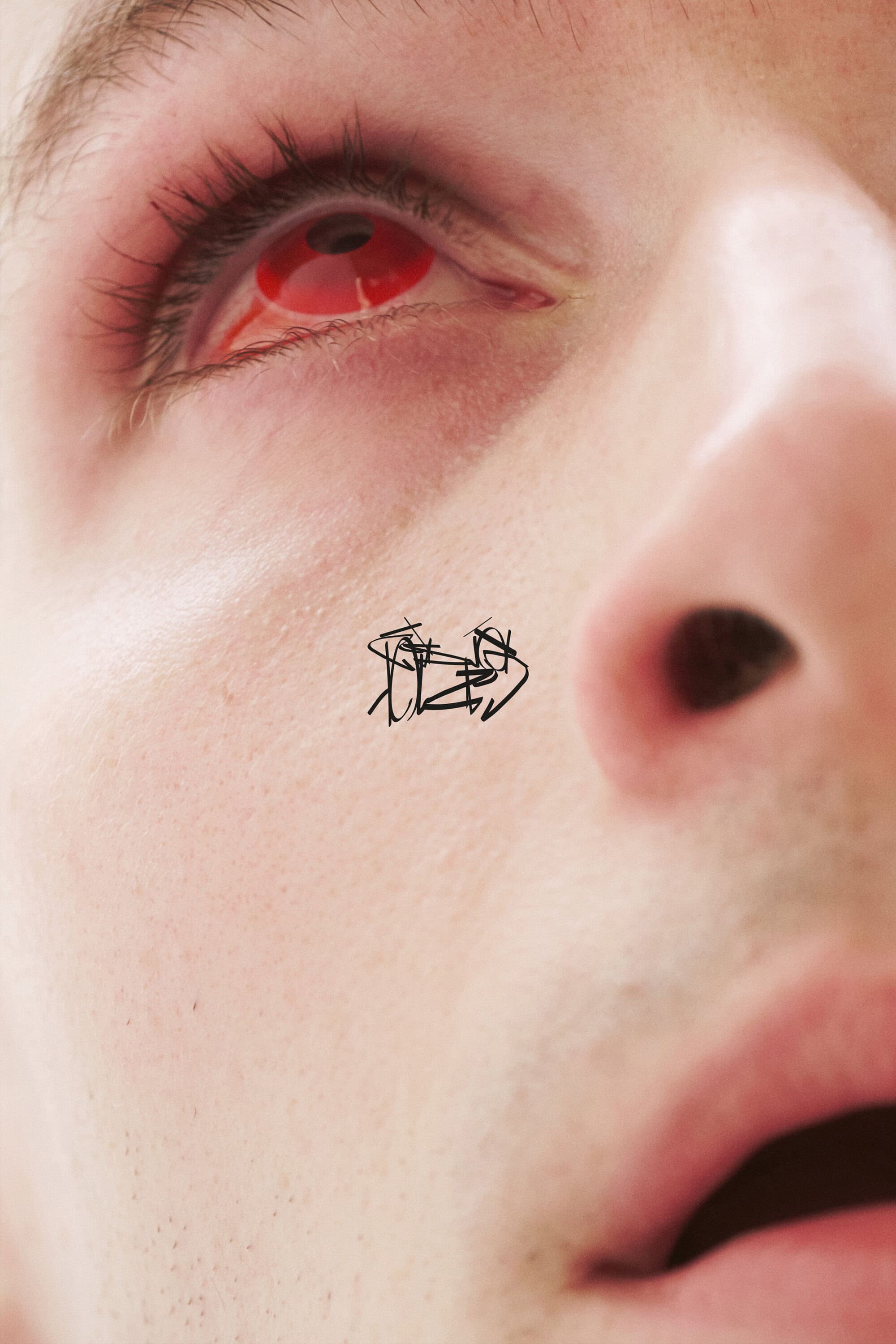 Extreme close-up portrait of a person's eye with a red contact lens and a small black scribble tattoo on the cheek.