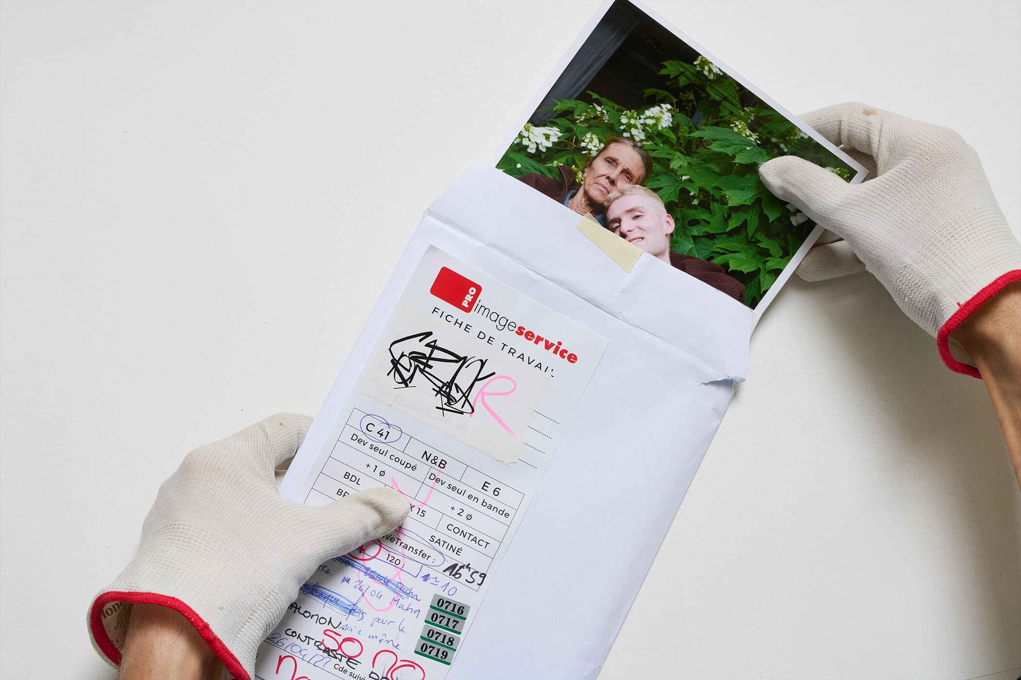 Gloved hands opening a white envelope containing a photograph of two people amidst green foliage, for GR10K.