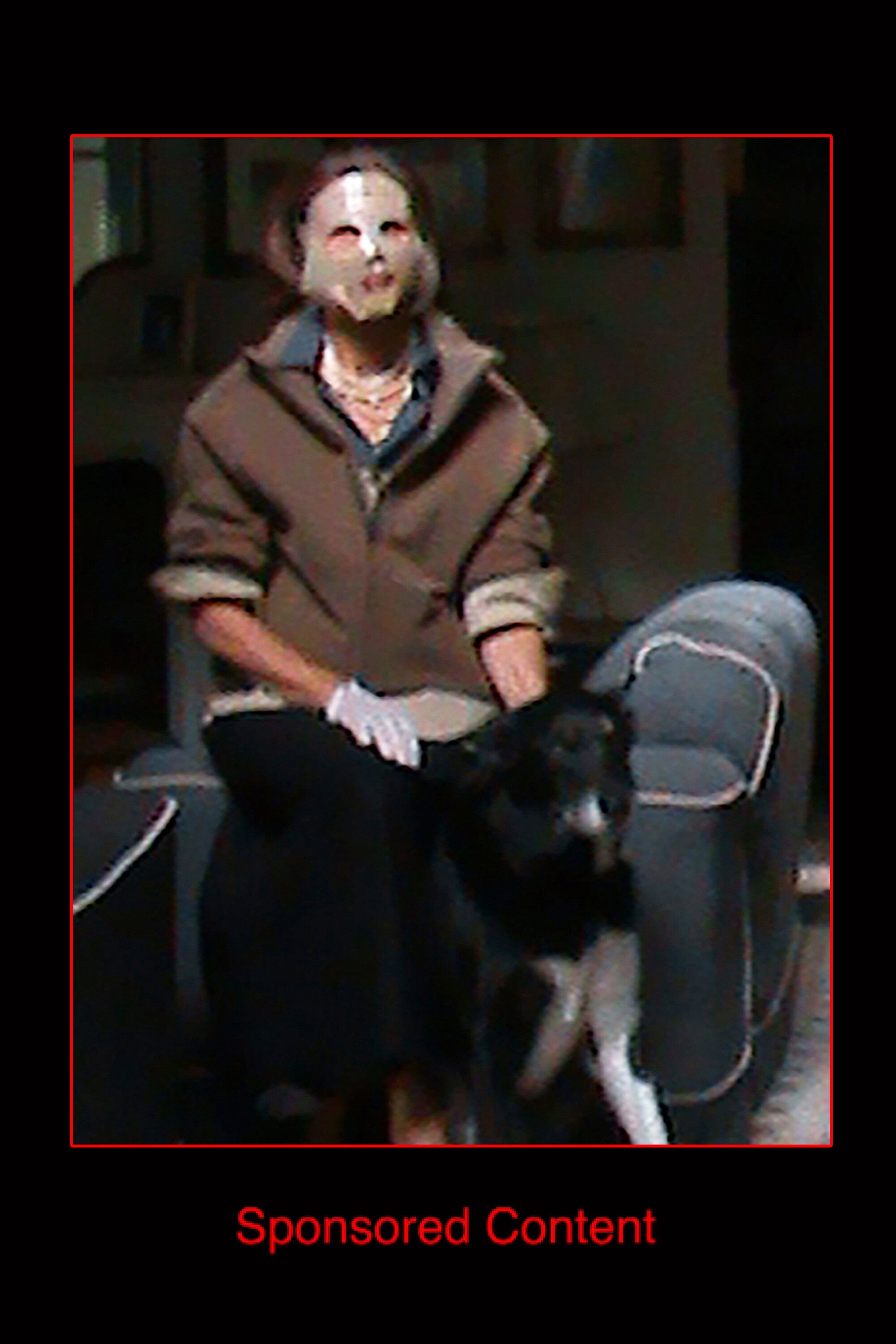 Person seated on a chair wearing a brown jacket and a face covering next to a dark dog in an indoor setting