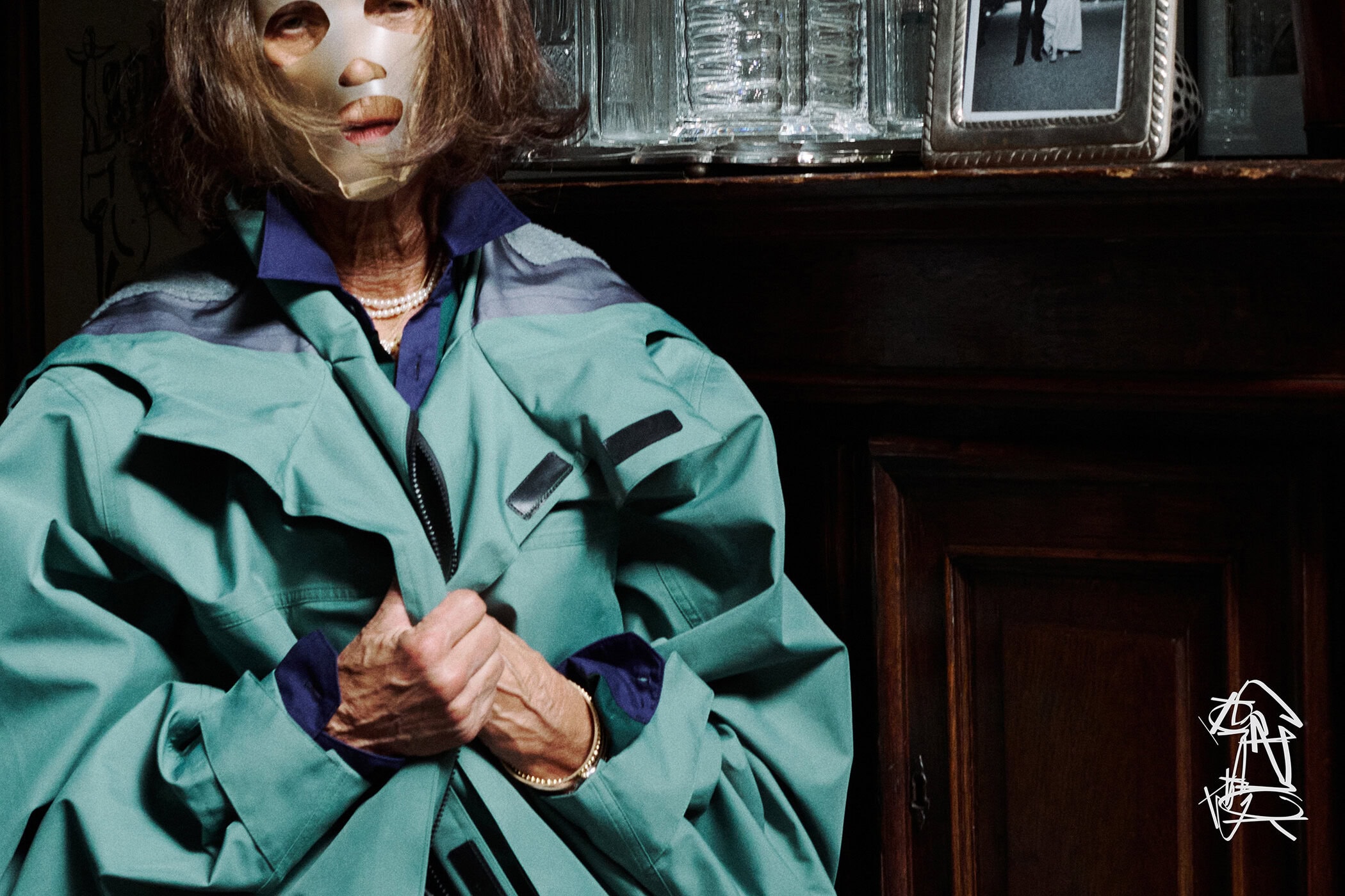 An elderly woman wearing a teal jacket and a metallic face mask indoors near a dark wooden cabinet.