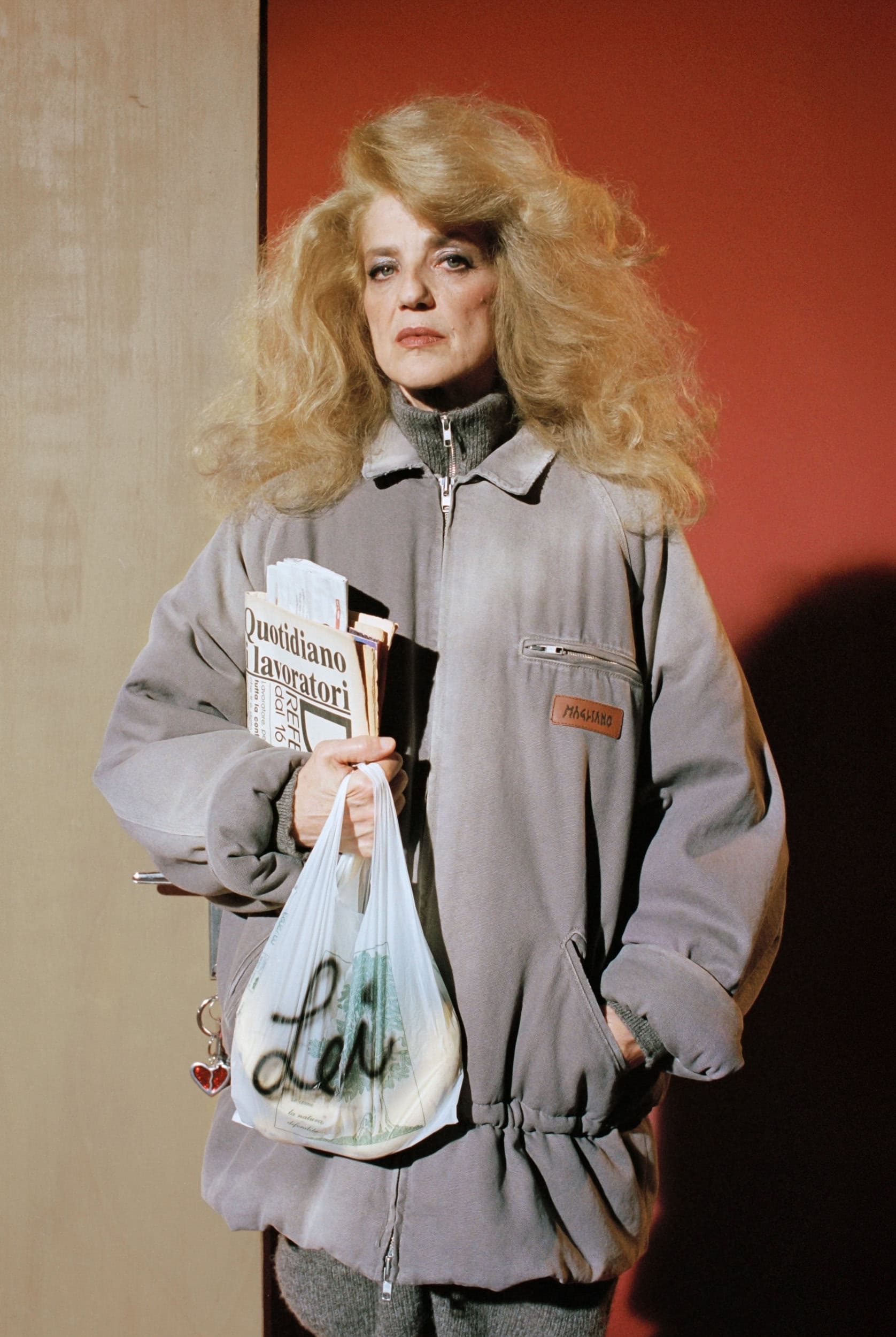 Woman with voluminous blonde hair holding a plastic shopping bag and newspapers wearing a grey jacket
