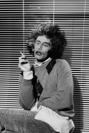 Black and white portrait of a person with curly hair holding a cigarette against window blinds