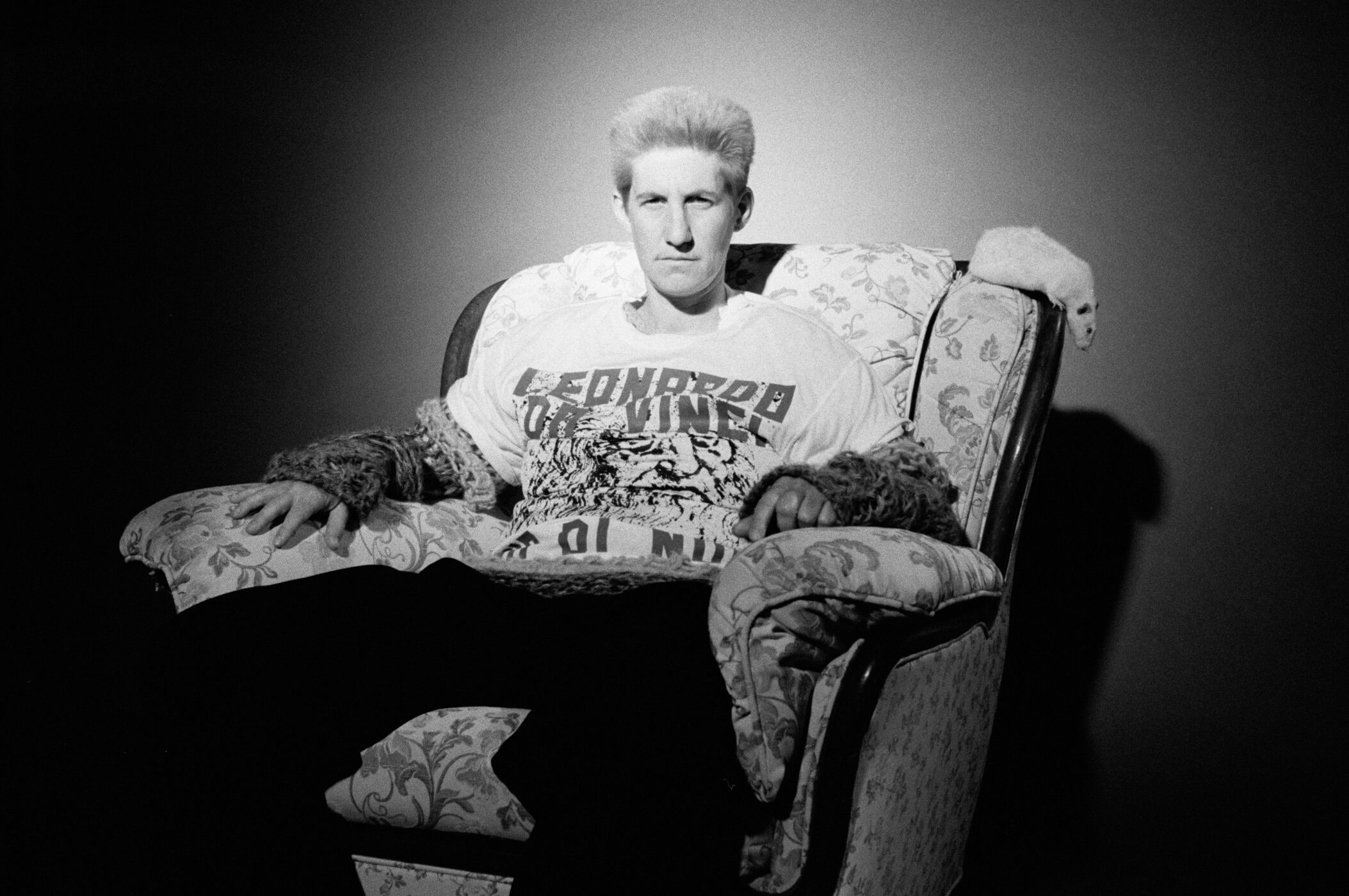 Monochrome portrait of a man with light hair seated in a patterned armchair wearing a printed t-shirt