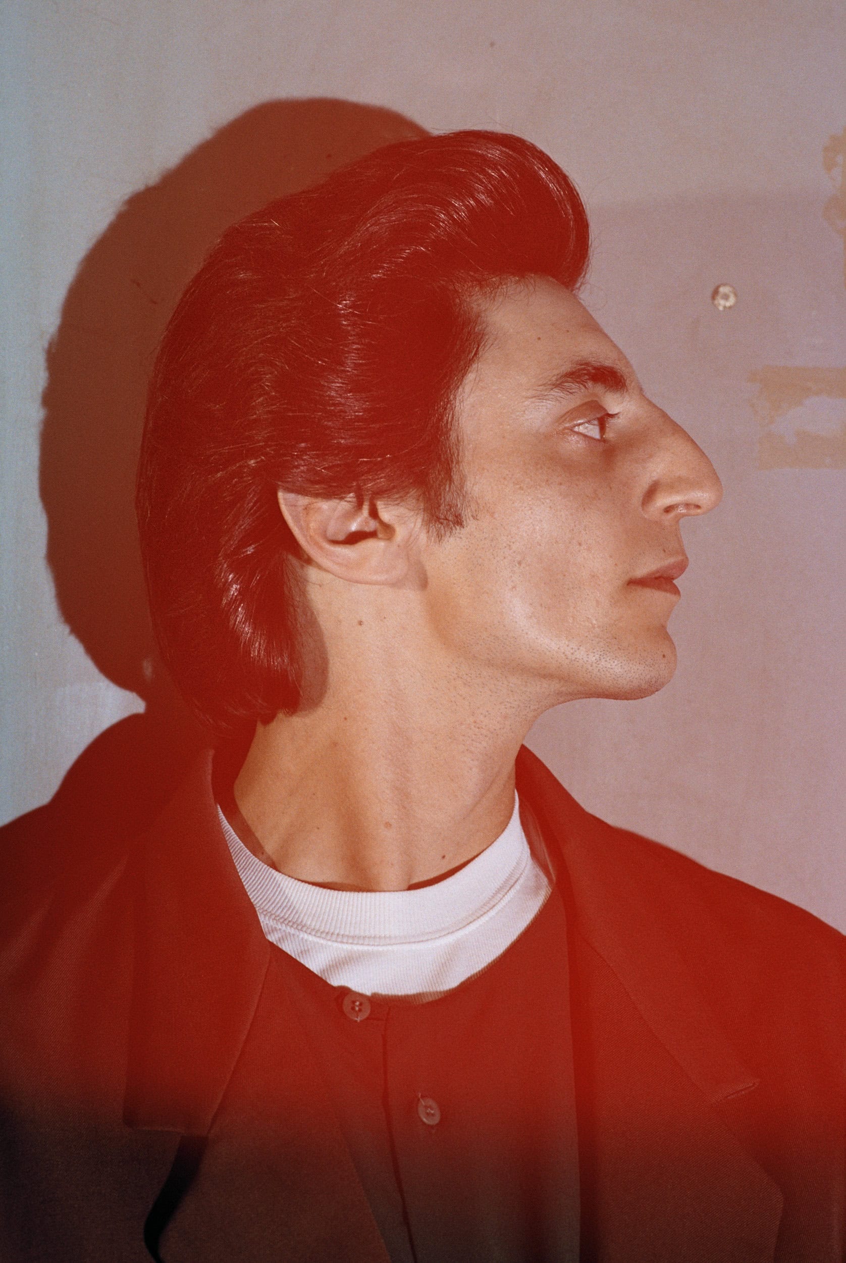 Profile portrait of a man with dark styled hair wearing a dark jacket over a white collared shirt against a wall.