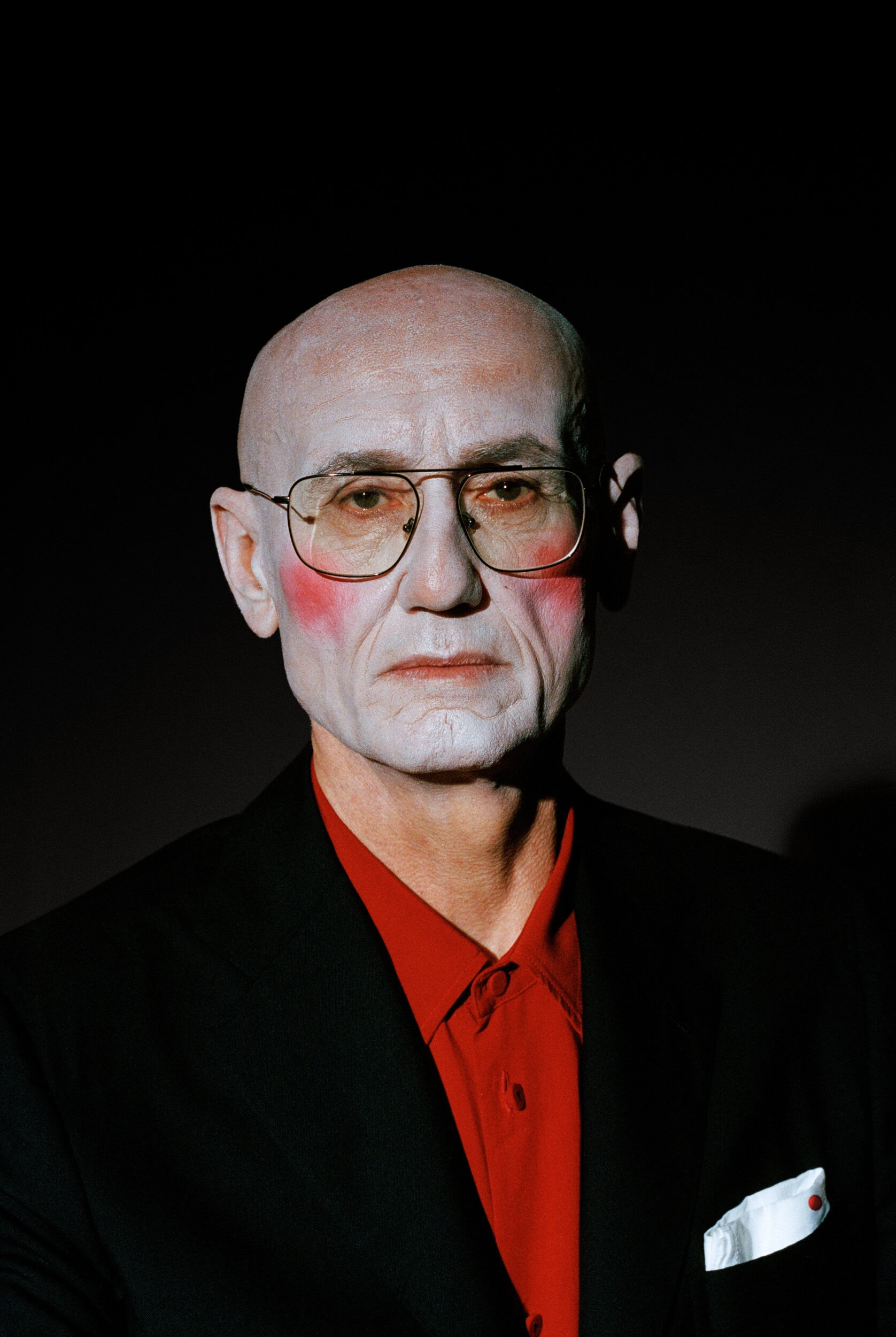Portrait of an elderly bald man wearing tinted aviator glasses, a red shirt, and a black blazer against a dark background.
