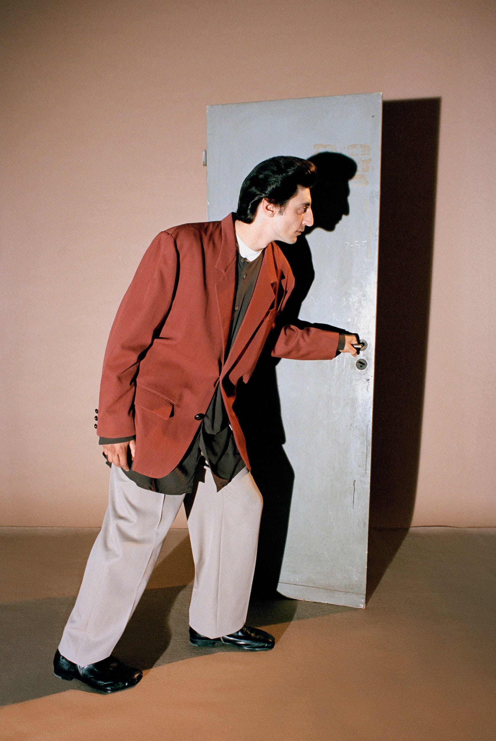 Man wearing a brown blazer and grey trousers opening a grey metal door set against a plain brownish studio background.