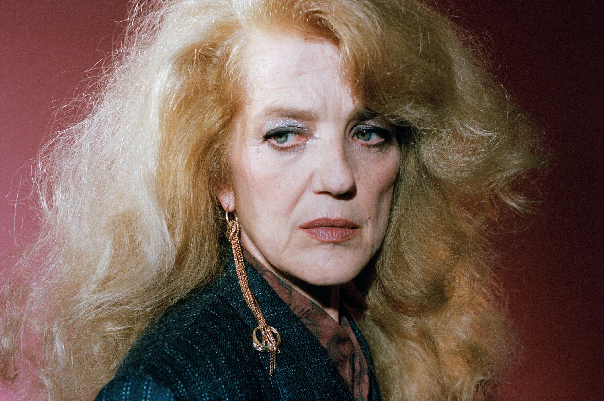 Portrait of an older woman with voluminous blonde hair wearing a dark textured blazer and long drop earrings against a red backdrop.