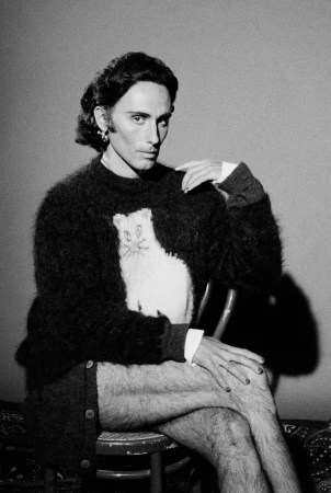 Portrait of a man seated on a chair wearing a black fluffy sweater with a white cat graphic for Magliano.