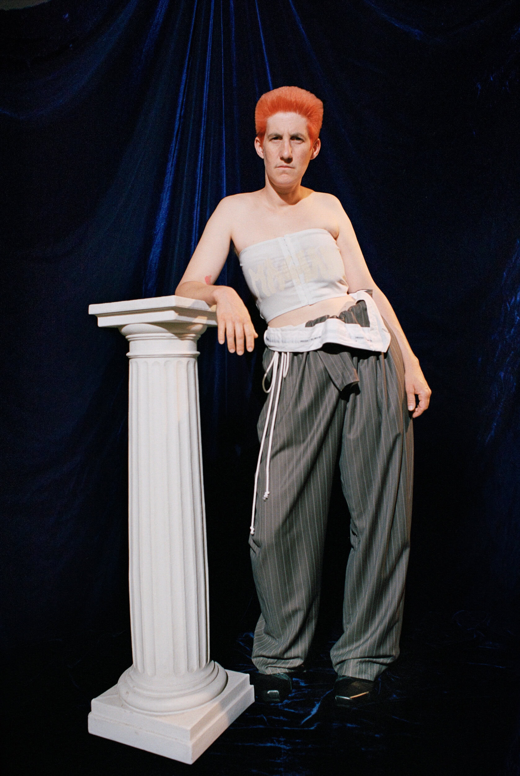 Person with orange hair wearing a white top and striped trousers leaning on a white classical column against dark blue fabric