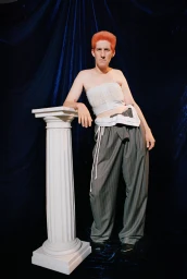 Person with orange hair wearing a white top and striped trousers leaning on a white classical column against dark blue fabric