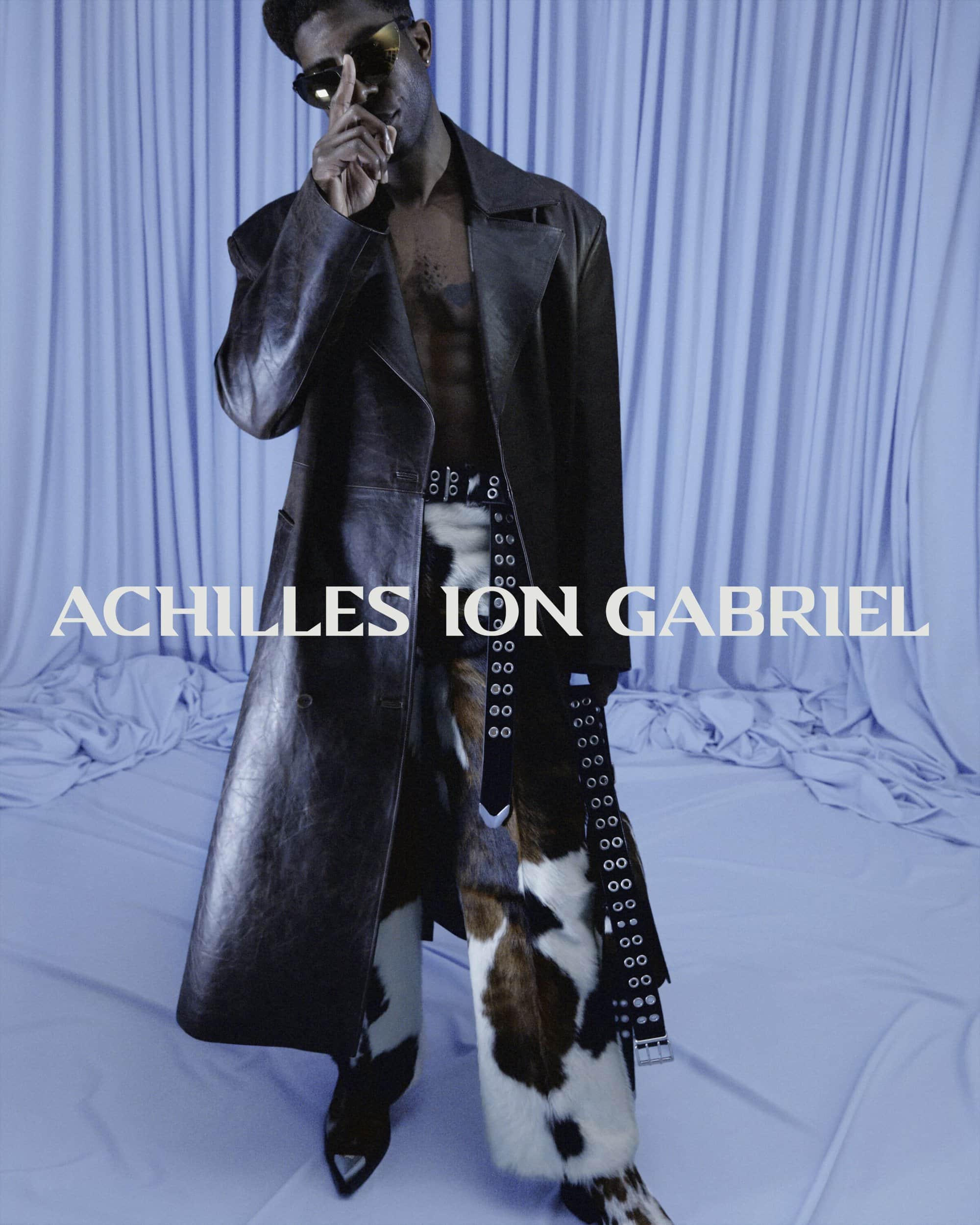 Man wearing a long dark coat and patterned trousers in front of draped blue fabric for Achilles Ion Gabriel