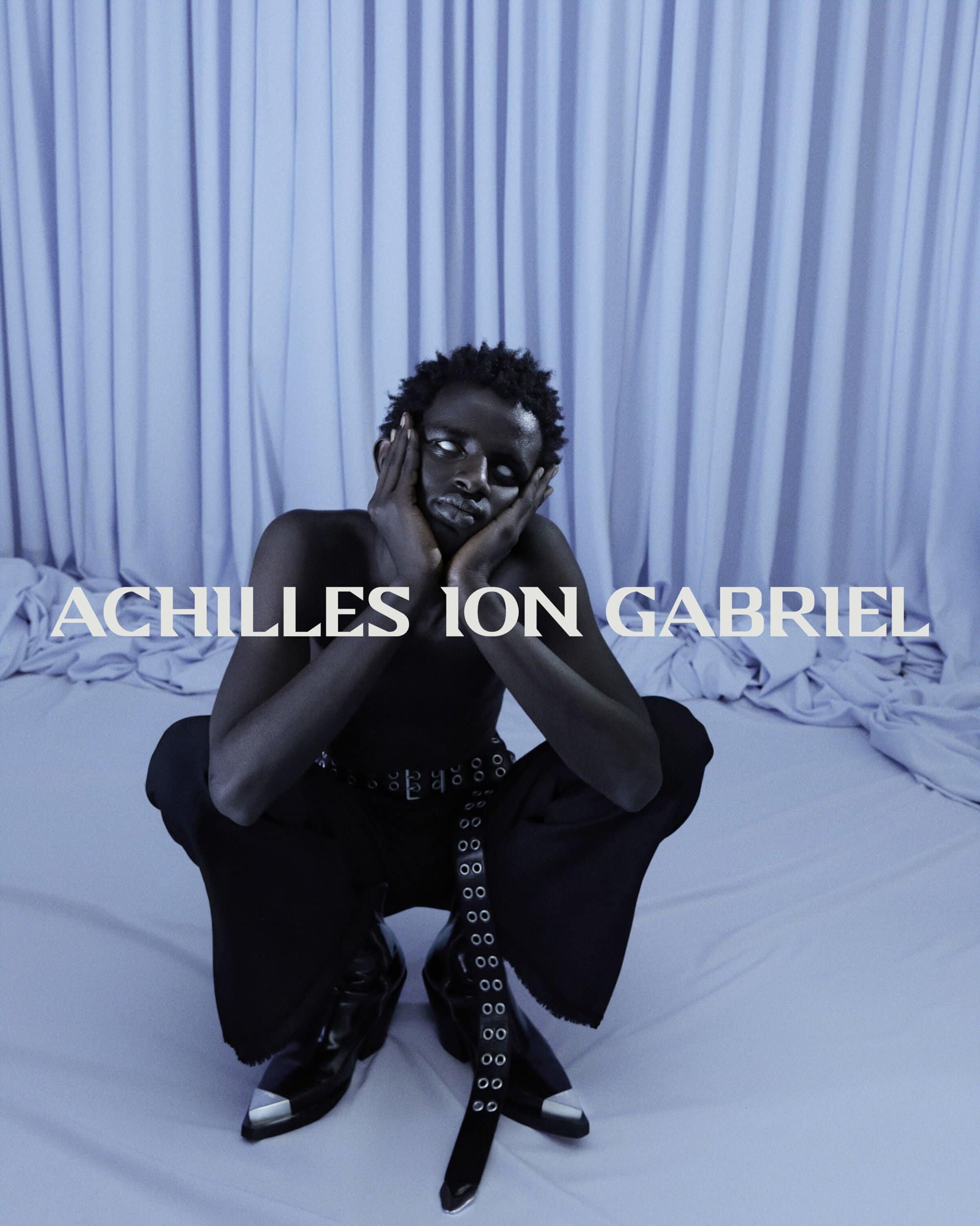Model squatting in black pants and metallic-toe boots against a pale draped backdrop for Achilles Ion Gabriel Fall 2024 ad campaign