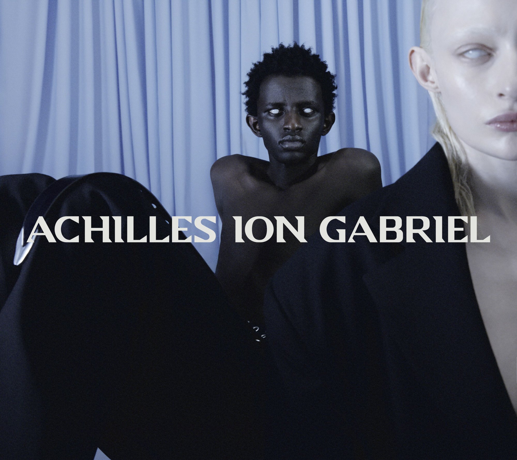 Two models posing against a draped pale blue background with overlaid white text reading ACHILLES ION GABRIEL.