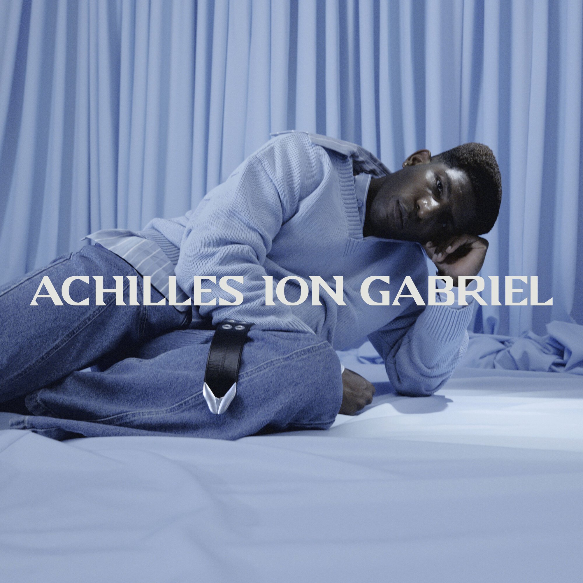 Model reclining on a blue draped surface wearing a light blue knit sweater and denim jeans for Achilles Ion Gabriel
