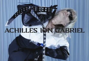 Model wearing dark sunglasses, white top, and black trousers holding a handbag and studded belt against a pale blue draped backdrop