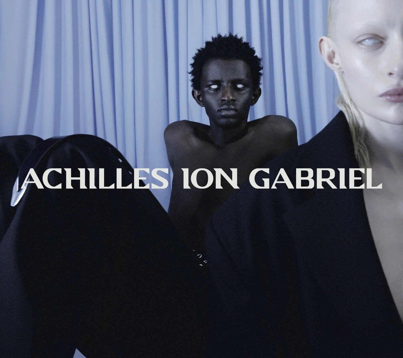 Two models posing against a draped pale blue background with overlaid white text reading ACHILLES ION GABRIEL.
