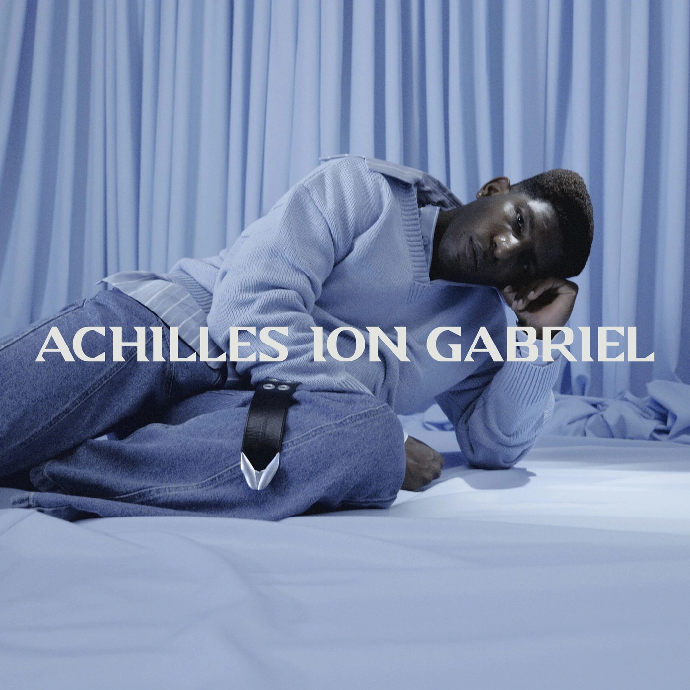 Model reclining on a blue draped surface wearing a light blue knit sweater and denim jeans for Achilles Ion Gabriel