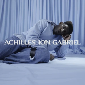 Model reclining on a blue draped surface wearing a light blue knit sweater and denim jeans for Achilles Ion Gabriel