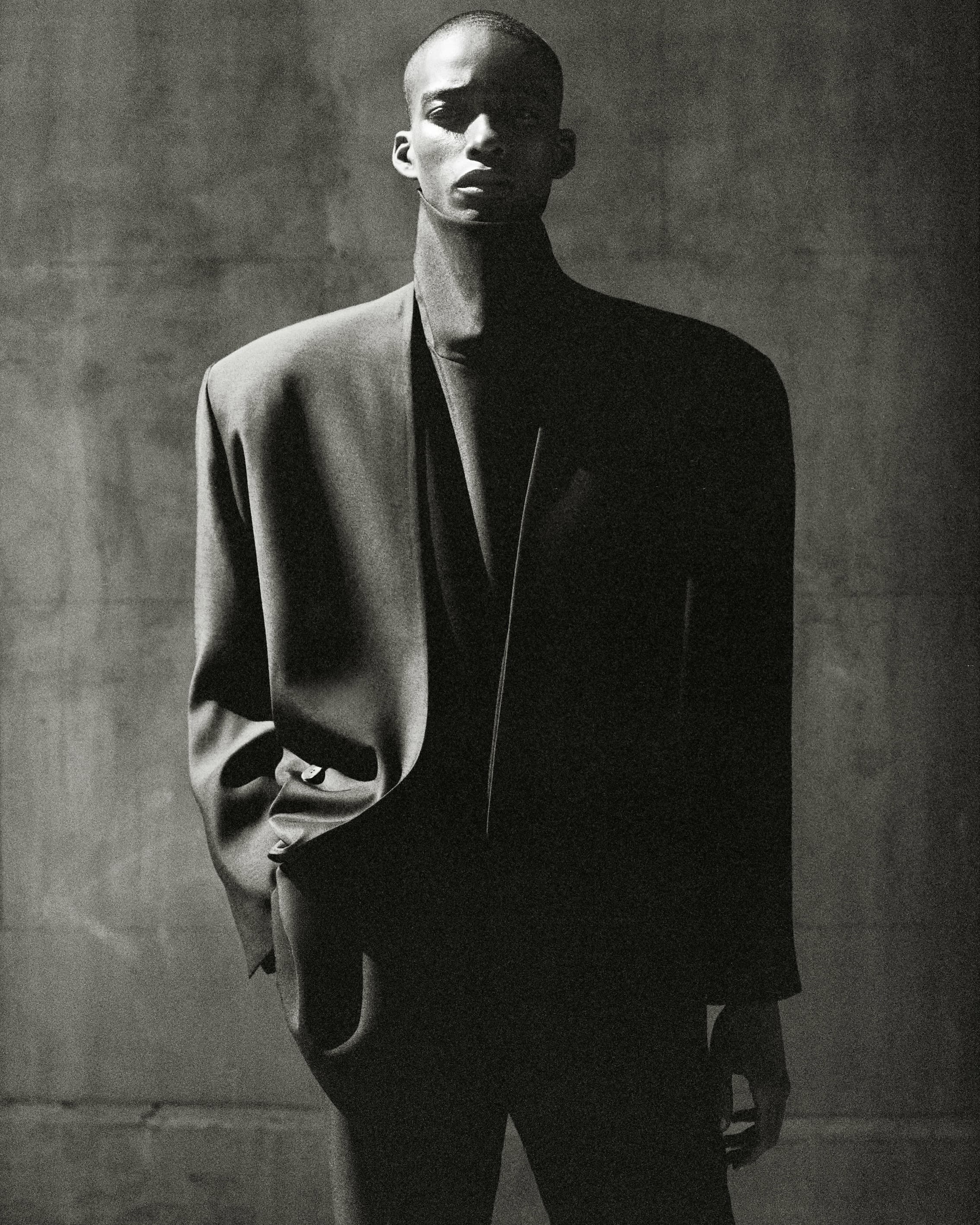 Black man wearing structured oversized black blazer against a dark textured wall in a monochrome portrait.