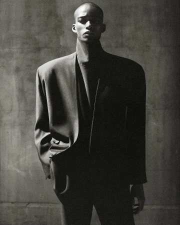 Black man wearing structured oversized black blazer against a dark textured wall in a monochrome portrait.