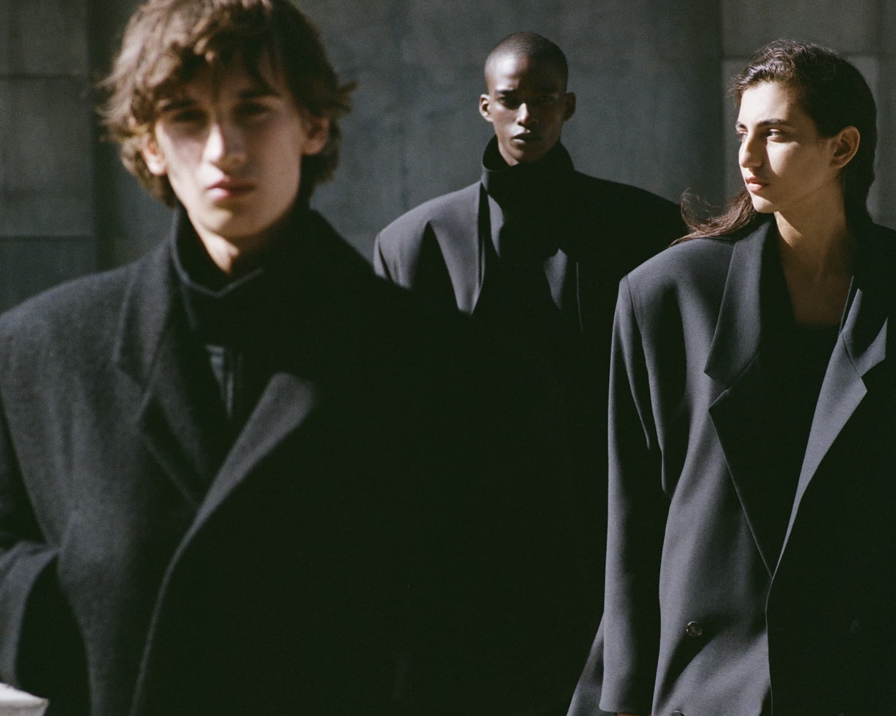Three models wearing structured black coats and blazers outdoors in natural sunlight
