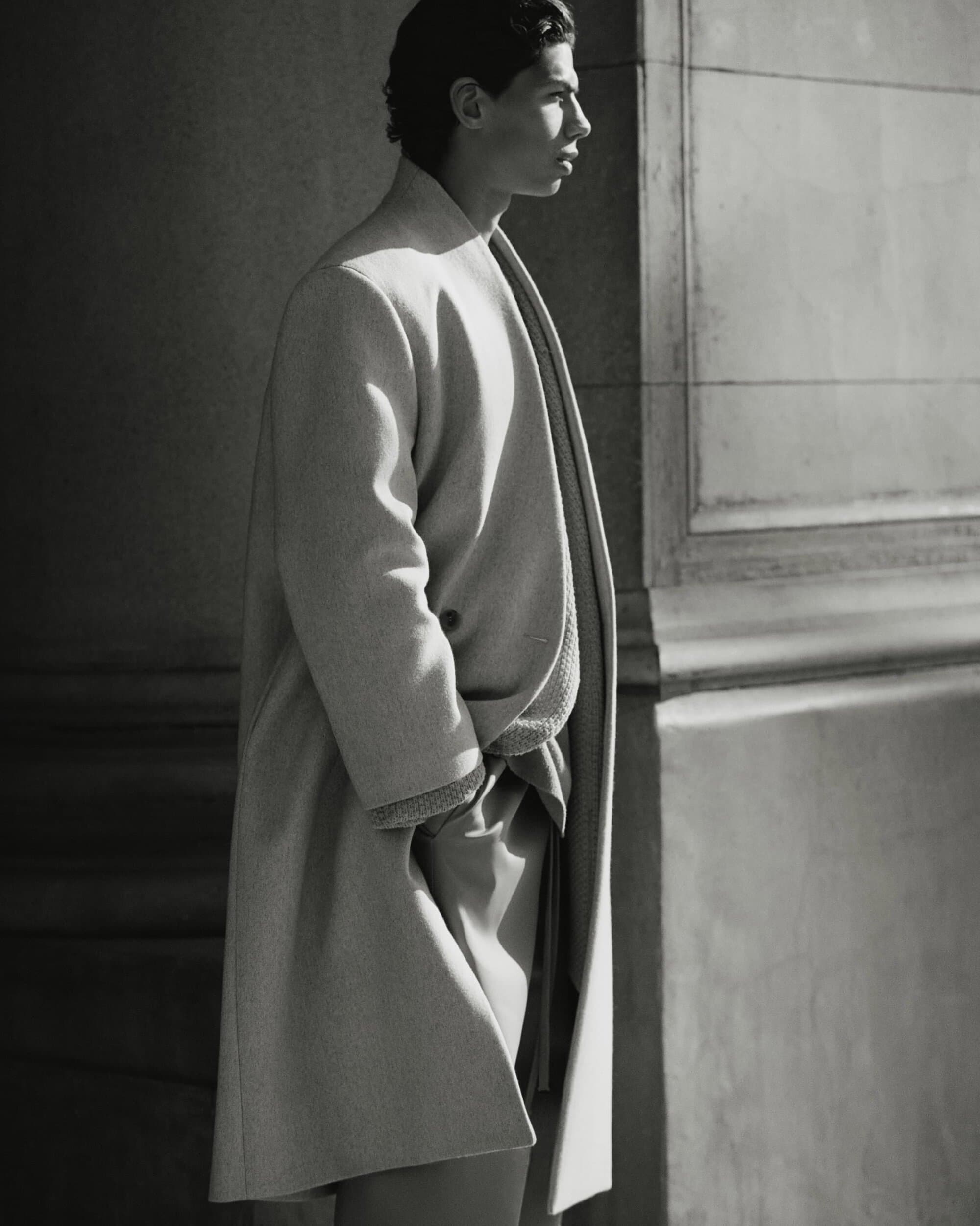 Man in long coat standing beside stone architecture in dramatic lighting